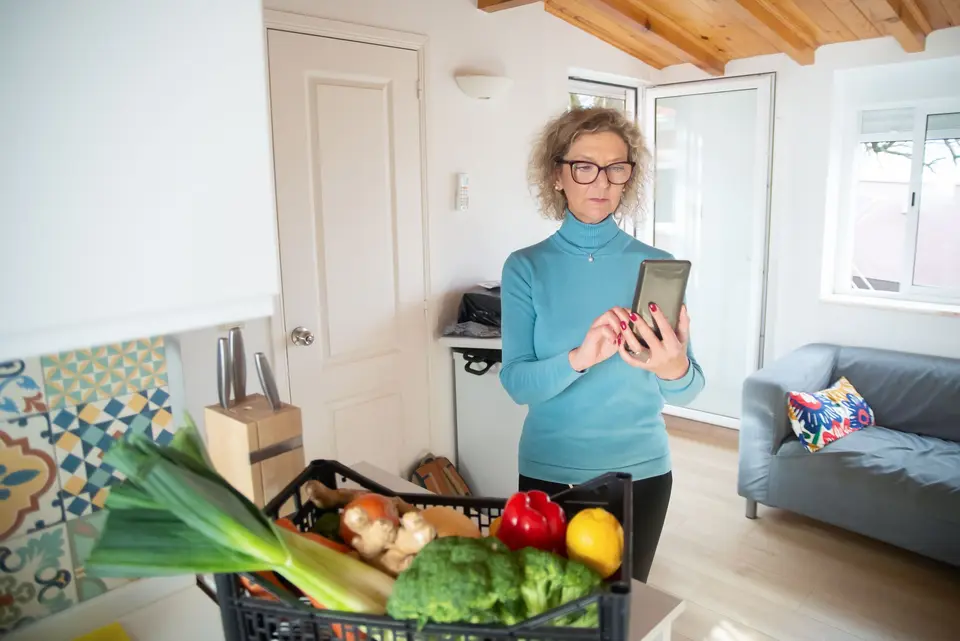 Een vrouw met bril staat in haar keuken en leest een bericht op haar telefoon